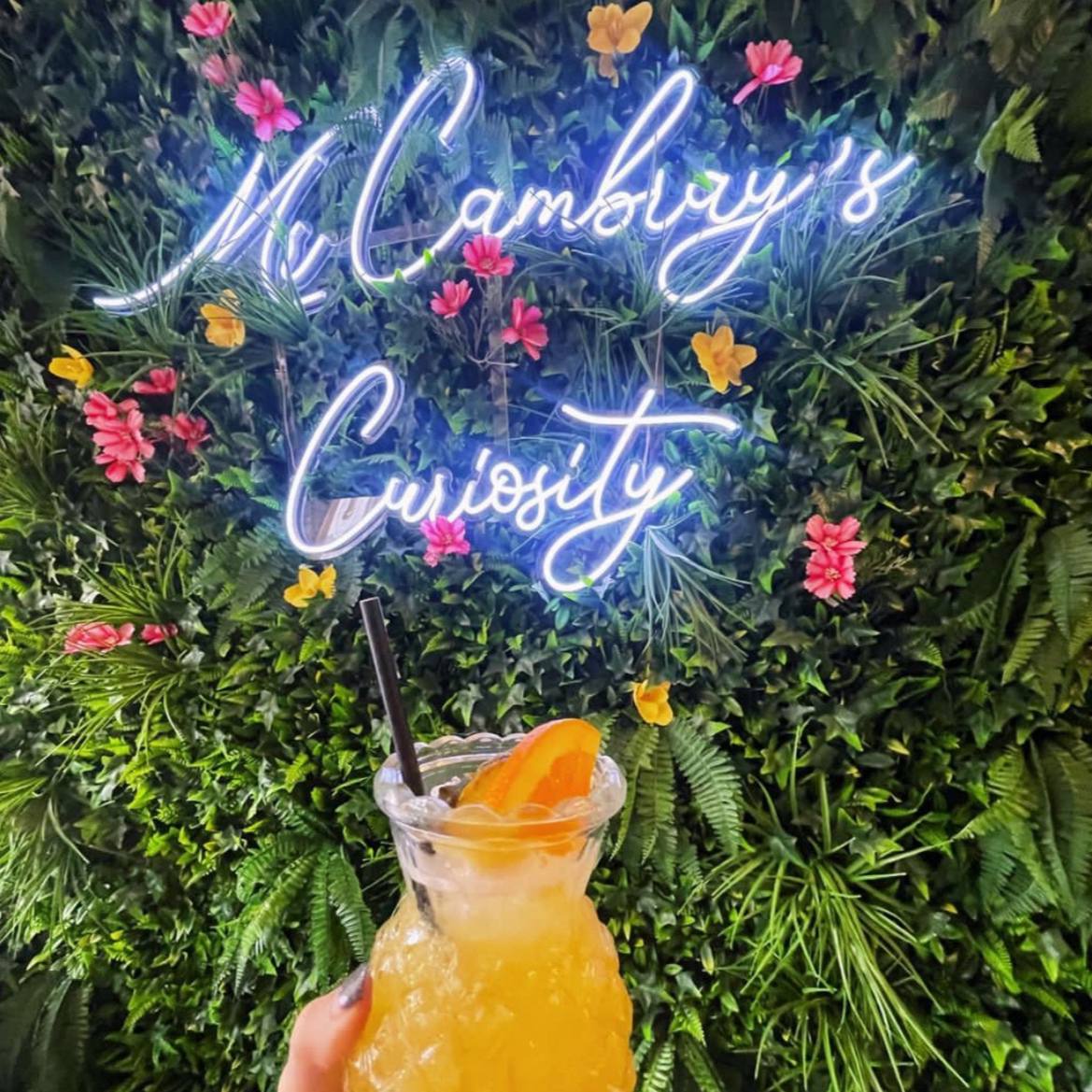 White script neon sign displayed on a colourful flower wall beside a cocktail