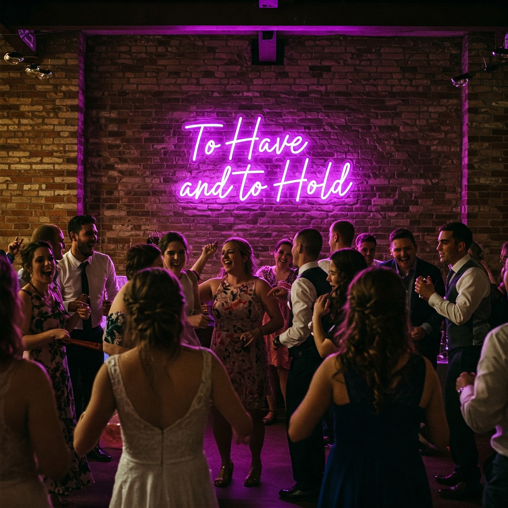 Purple “To Have and To Hold” neon sign on a brick wall above a wedding dancefloor with guests celebrating